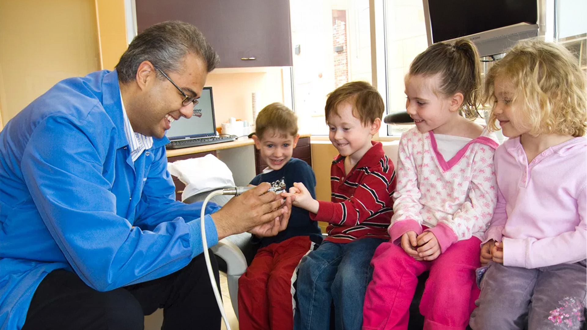 In the image, a man wearing a blue coat is kneeling down next to three children who are sitting on the floor, all looking at something in his hands.