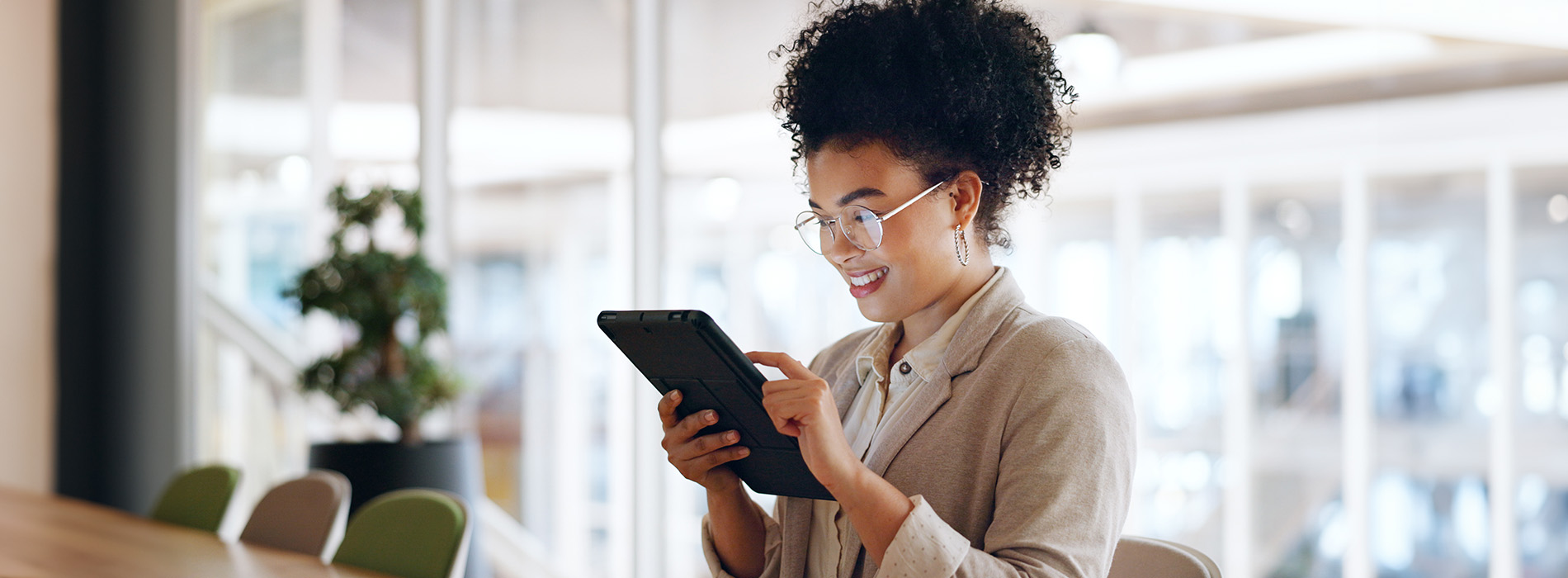 A woman standing in a modern indoor setting, engrossed in her phone, which she holds in both hands.