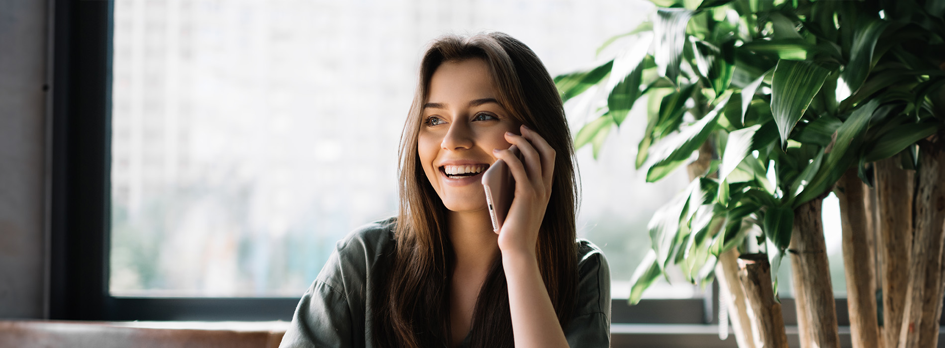 A young woman sitting at a table with a potted plant behind her, smiling while talking on her phone.