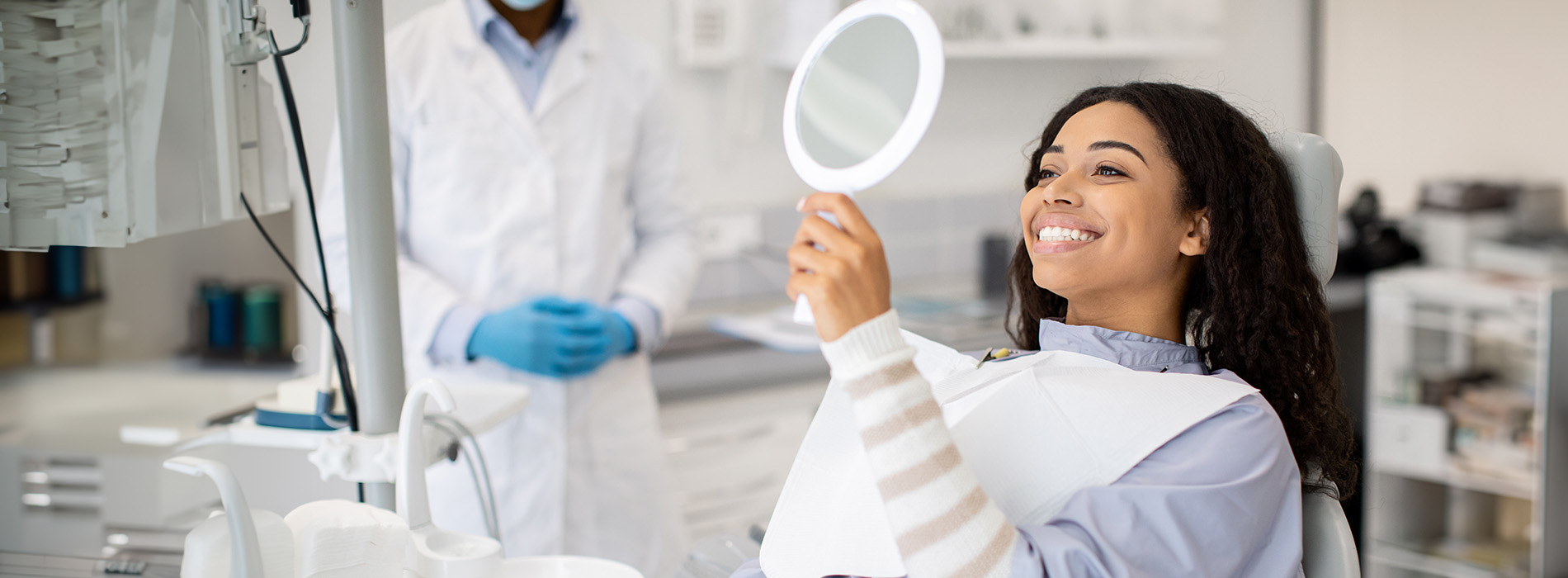 A woman with a bright smile sits at a dental station, surrounded by medical equipment and a dental professional.