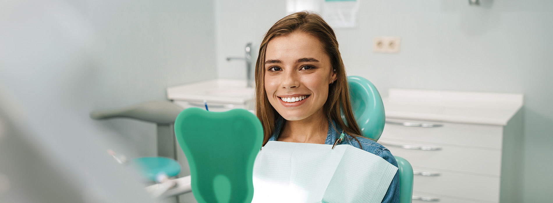 A young woman wearing a blue top sits at a dental chair with a smile on her face, looking towards the camera.
