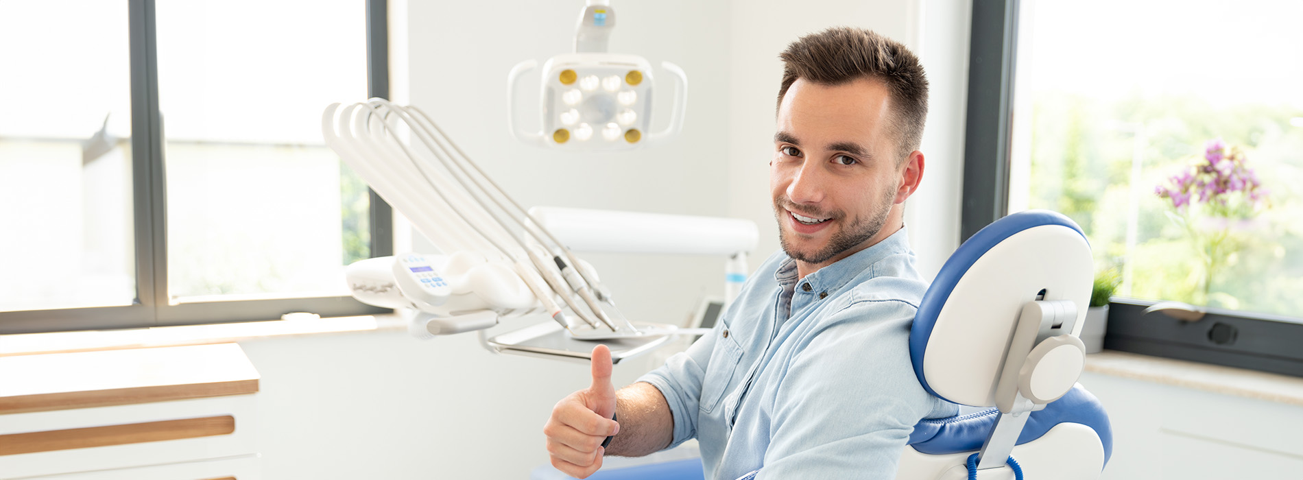 The image features a man sitting at a dental chair with a smile on his face, holding up a thumbs-up gesture.