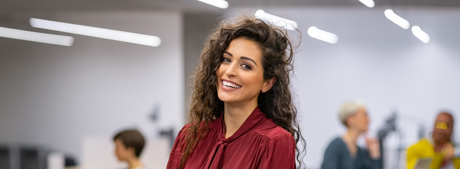 A young woman with long hair wearing a red top and standing in an indoor setting with other people in the background.