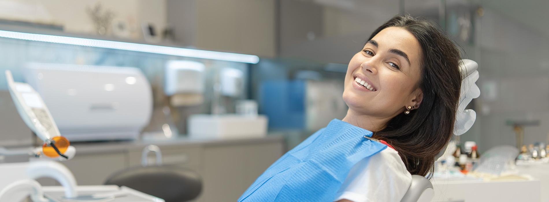 The image shows a smiling woman wearing blue scrubs, sitting in a dental chair with her eyes closed, likely in a dental office setting.