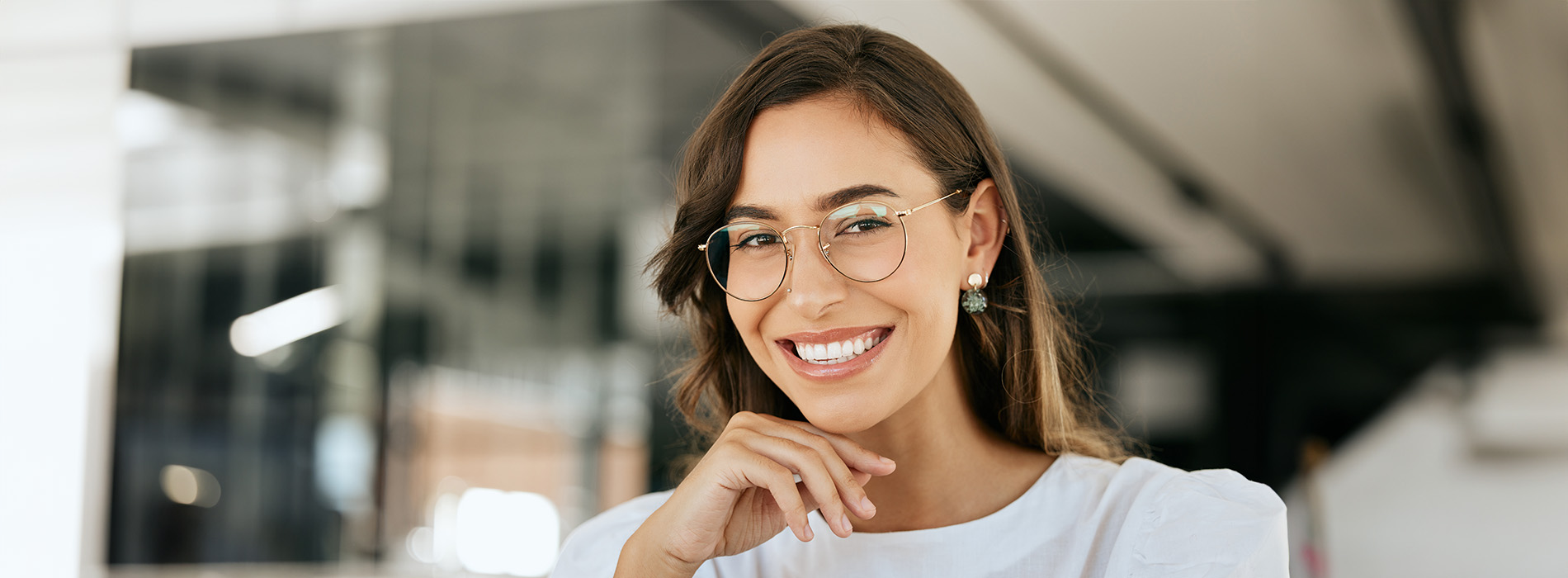 The image shows a woman smiling at the camera with her hands on her hips, wearing glasses and a white top, against a blurred background that includes an interior space with a modern design.