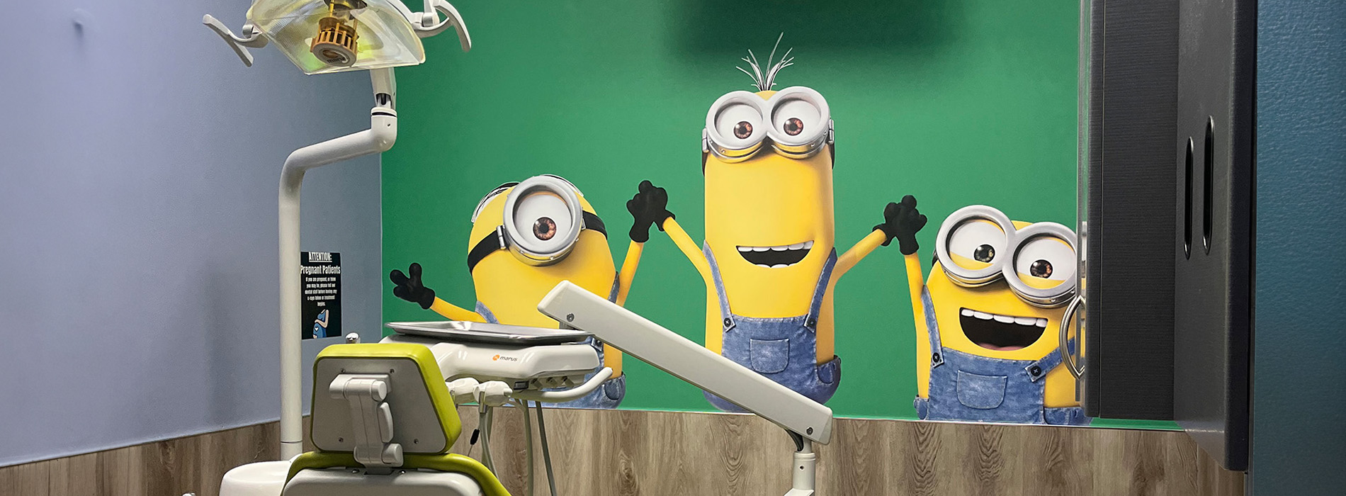 The image shows a room with a green background featuring three Minions characters on the wall, each holding up their hands in excitement.