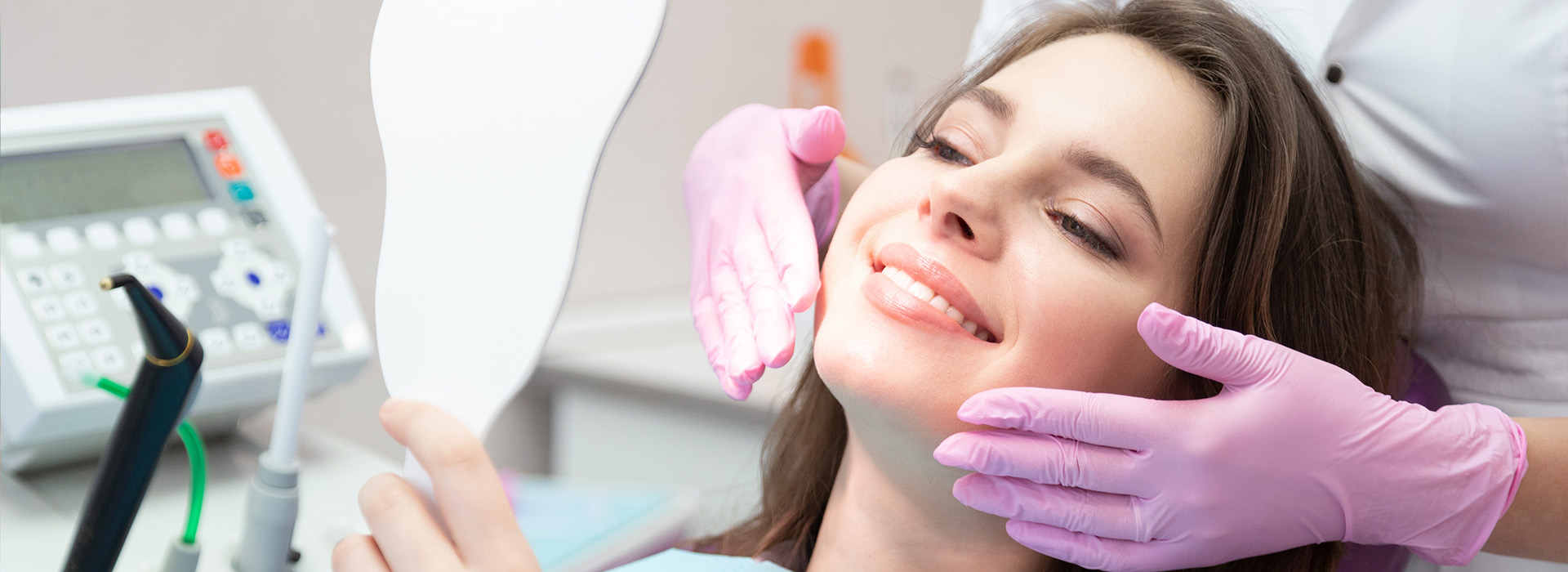A woman sitting in a dental chair with a hygienist working on her teeth, both wearing protective gear, against a blurred background featuring medical equipment.