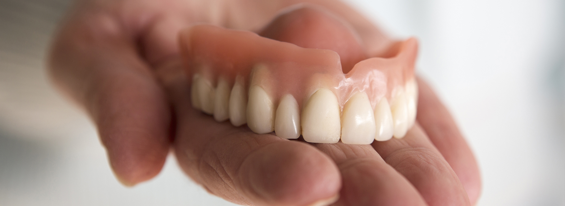 The image shows a hand holding an open mouth with a set of dentures against a blurred background.