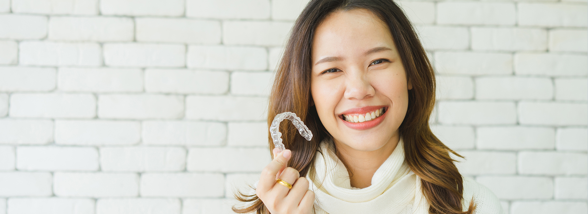 A woman with a toothy grin holds a toothbrush up to her mouth against a white brick wall backdrop.
