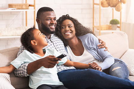 A family of three sits on a couch, with two adults holding a child, smiling at the camera.