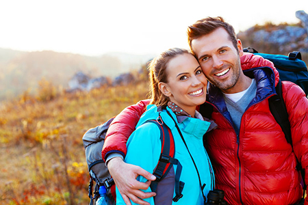 The image features a man and woman posing together outdoors, both with backpacks, smiling at the camera against a scenic background.