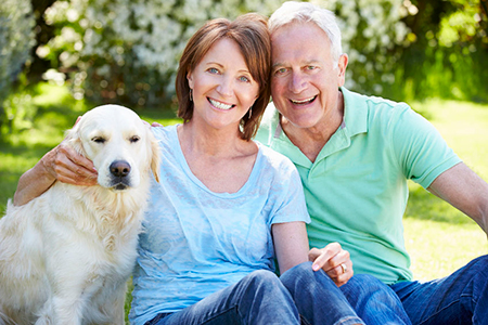 The image depicts an elderly couple sitting outdoors with their golden retriever dog, all smiling and enjoying a sunny day.