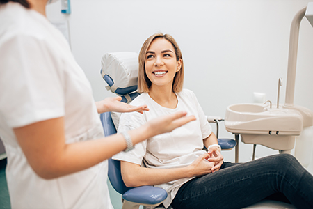 The image shows a woman seated on a dental chair with a smile, being attended by two dental professionals who appear to be assisting her during a dental appointment.
