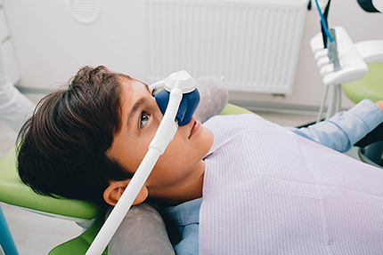 The image shows a person sitting in a dental chair with their eyes closed, wearing a surgical mask, and having their teeth cleaned by a dentist.